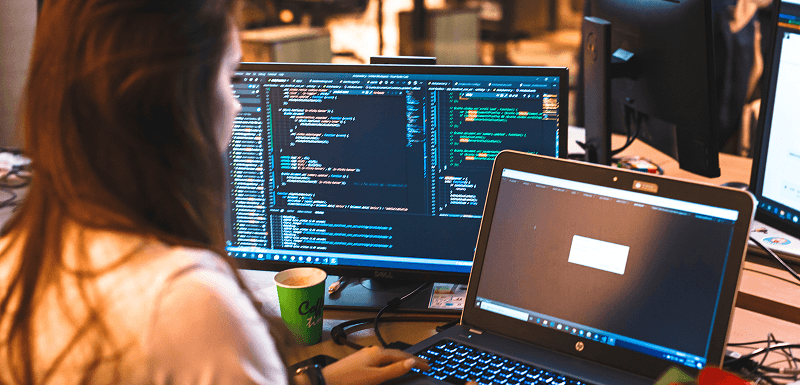 Woman working on a laptop beside monitors displaying computer code