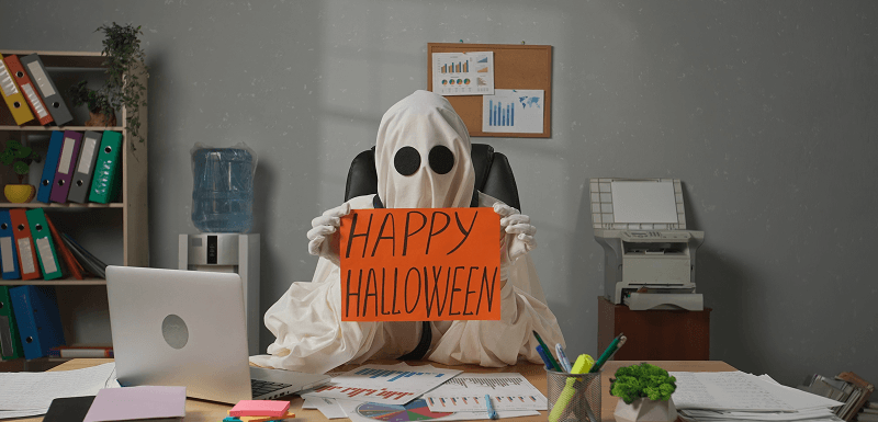 Person dressed as a ghost sits at an office desk holding a Happy Halloween sign