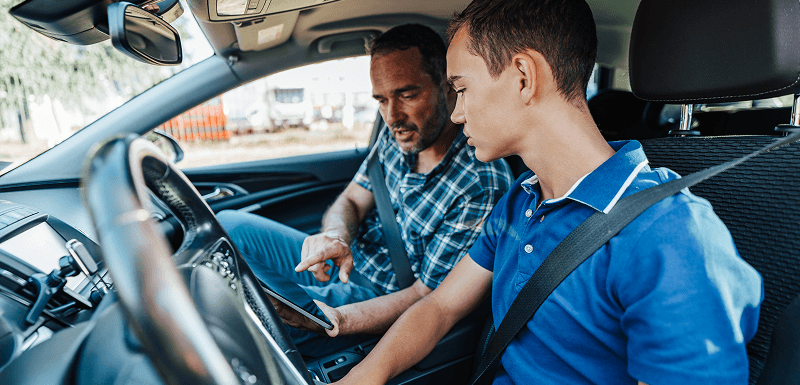 Two belted men in a car look at a tablet, with one pointing